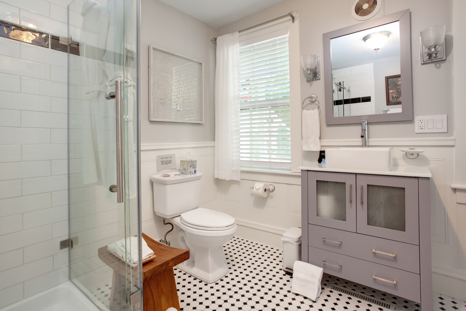 Bathroom with glass enclosed shower, toilet, gray cabinet with matching mirror, black and white tiled floor
