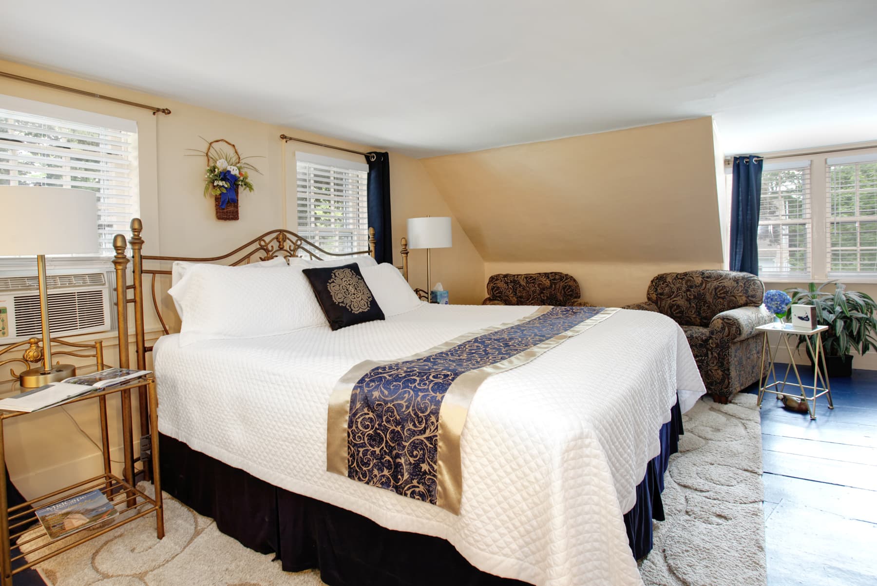 View of bed with white and navy bedding, two comfy upholstered chairs against far wall, nightstands with lamps, plush neutral area rug