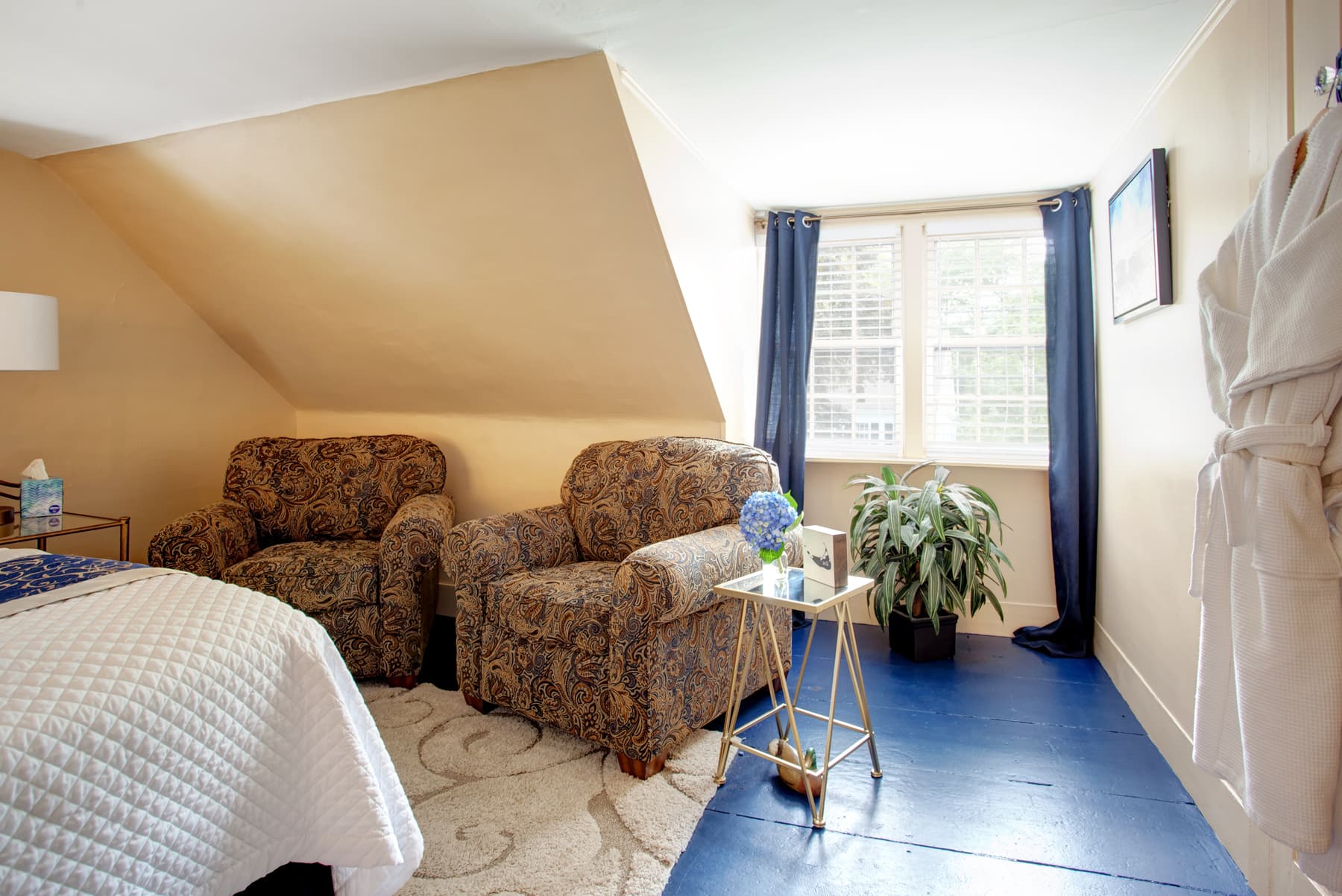 Sitting area to the side of bed with two comfy upholstered chairs, side table, bright sunlit window with navy curtains, potted plant under window