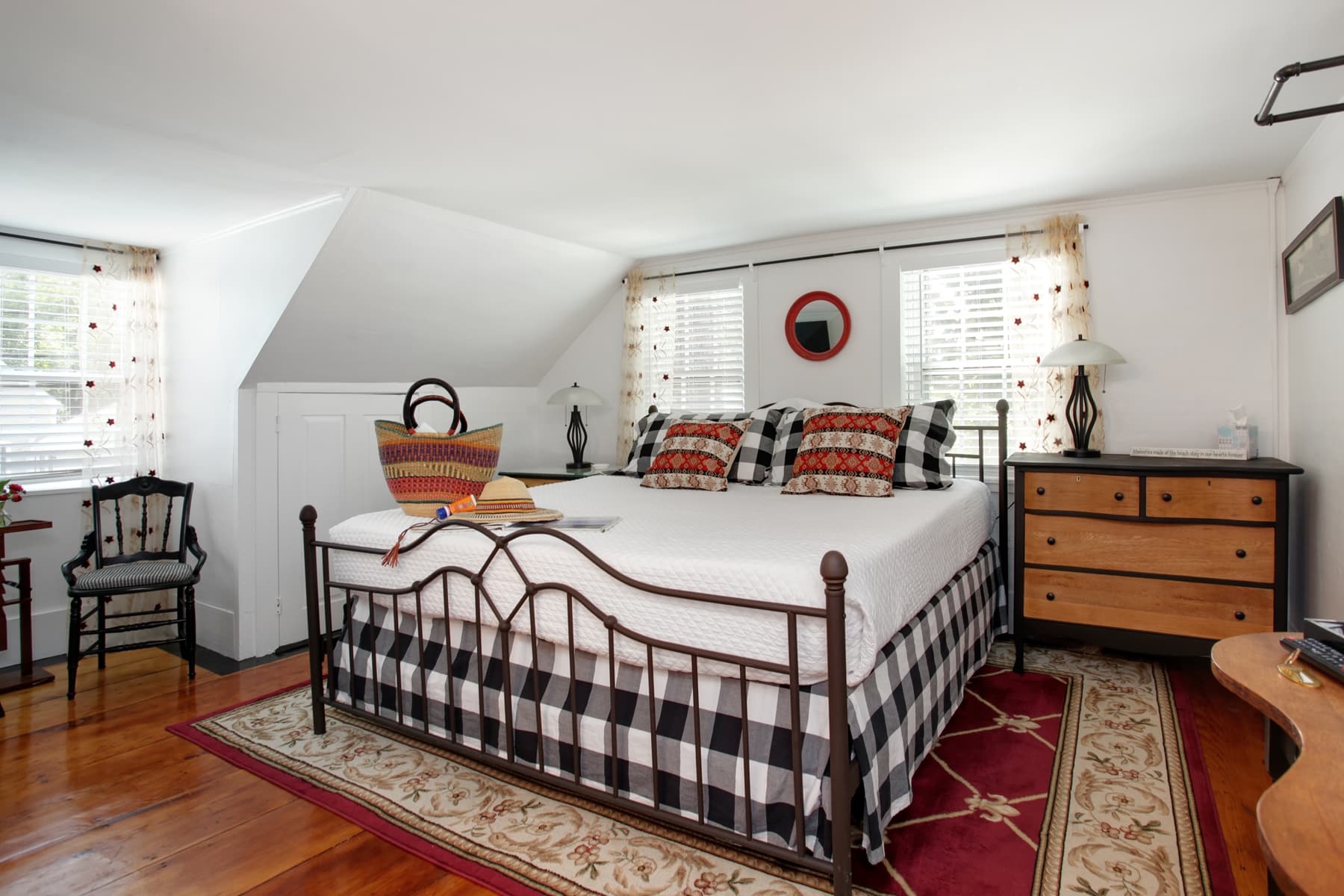 View of bed with black and white plaid bedding, natural wood and black painted bureau, and red and cream floral area rug