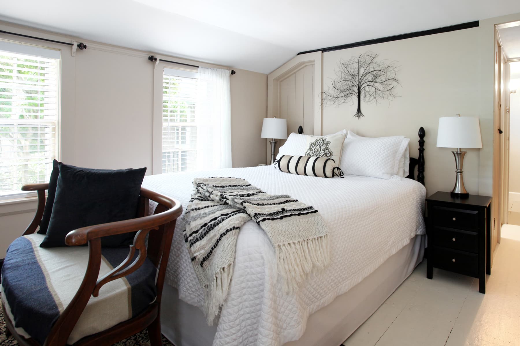 View of bed with white quilted bed cover, black painted wooden nightstand with lamp, and chair at foot of bed