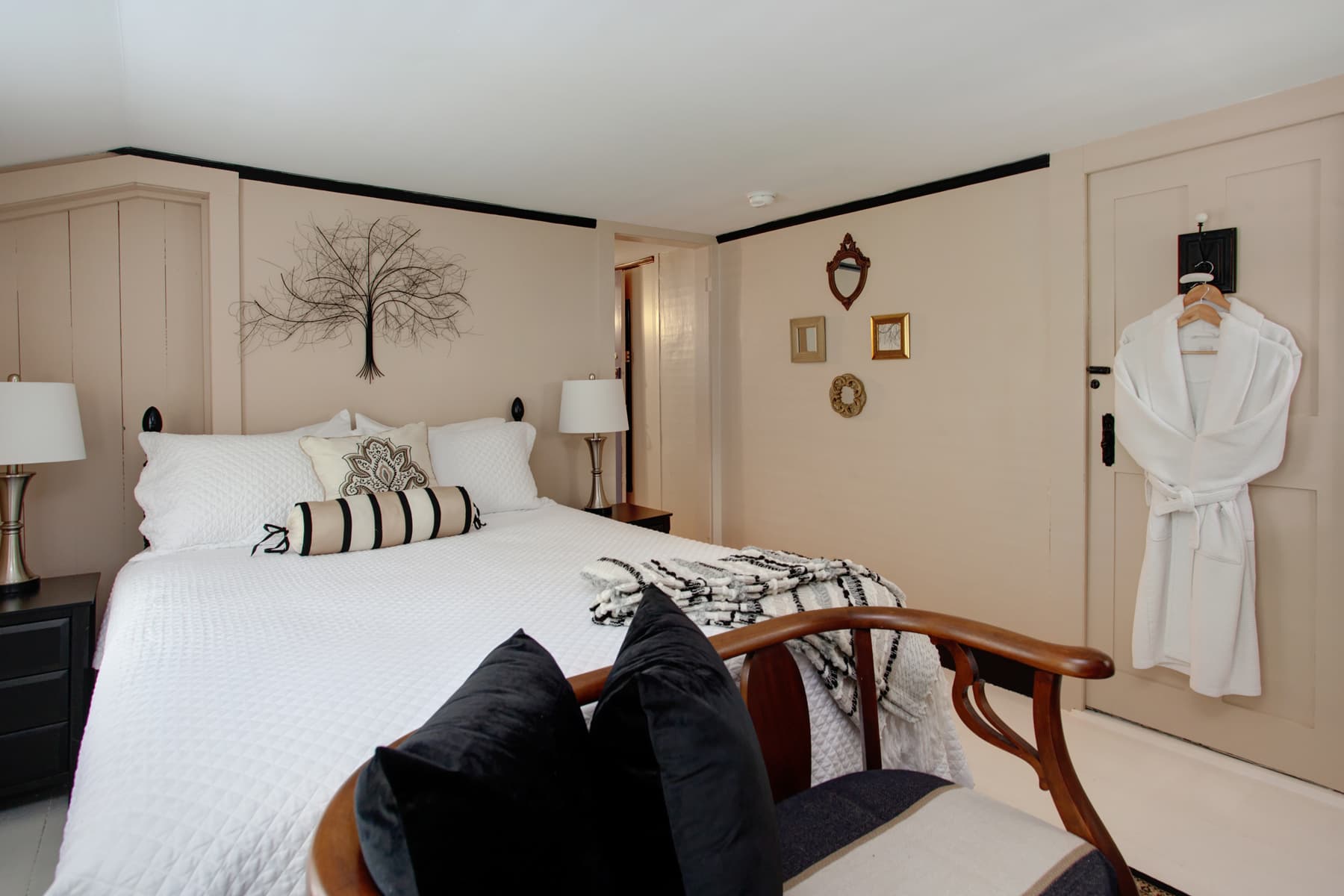 Bedroom with bed topped with white quilted bedcover, nightstands with matching lamps, decorative tree wall hanging, plush white robe hanging on wall, and wooden chair with cushions at foot of bed