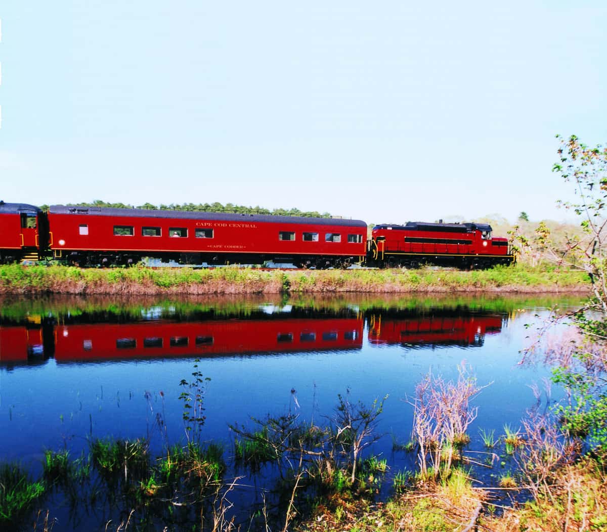 A red train travels alongside a calm body of water, reflecting its image.