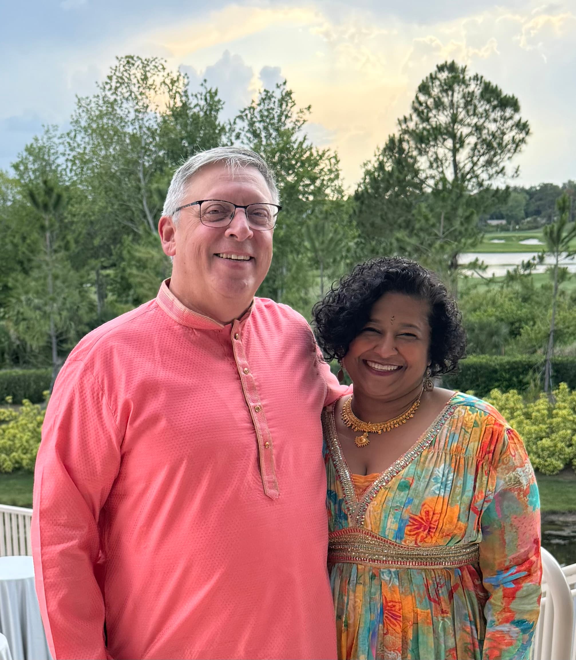 A smiling couple poses together outdoors, dressed in vibrant traditional attire against a scenic backdrop.