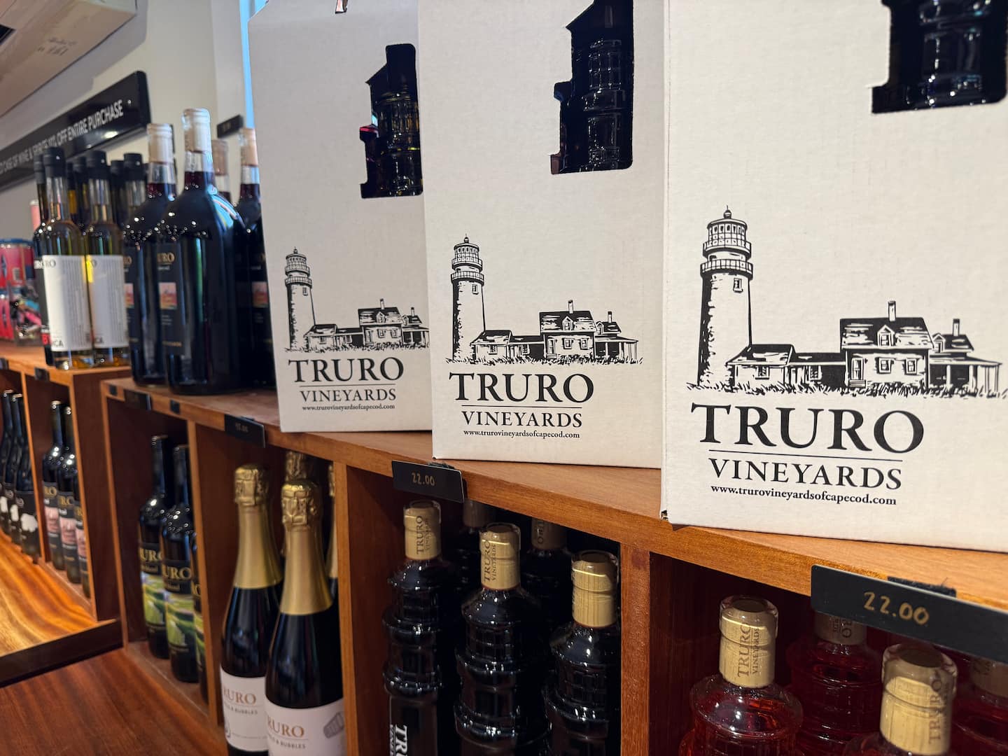 Wine boxes with the Truro Vineyards logo displayed on a wooden shelf among various bottles.