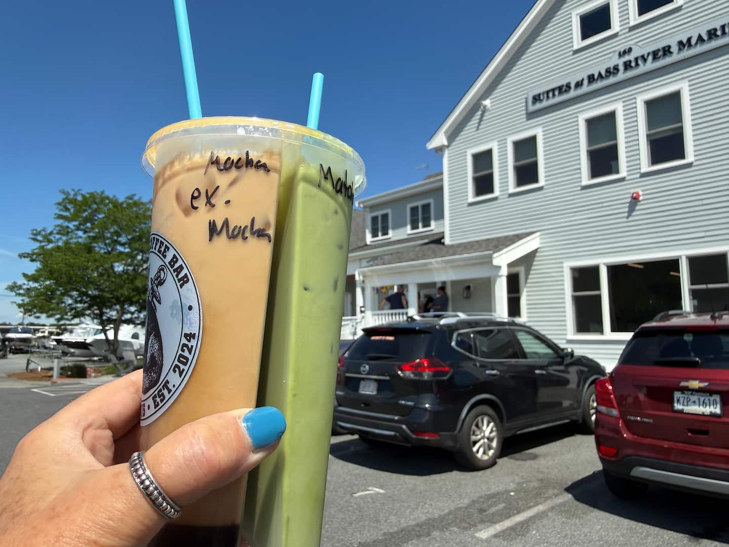 A hand holds two iced drinks, one mocha and one green, in front of a building labeled "Butters & Bass River Marina."