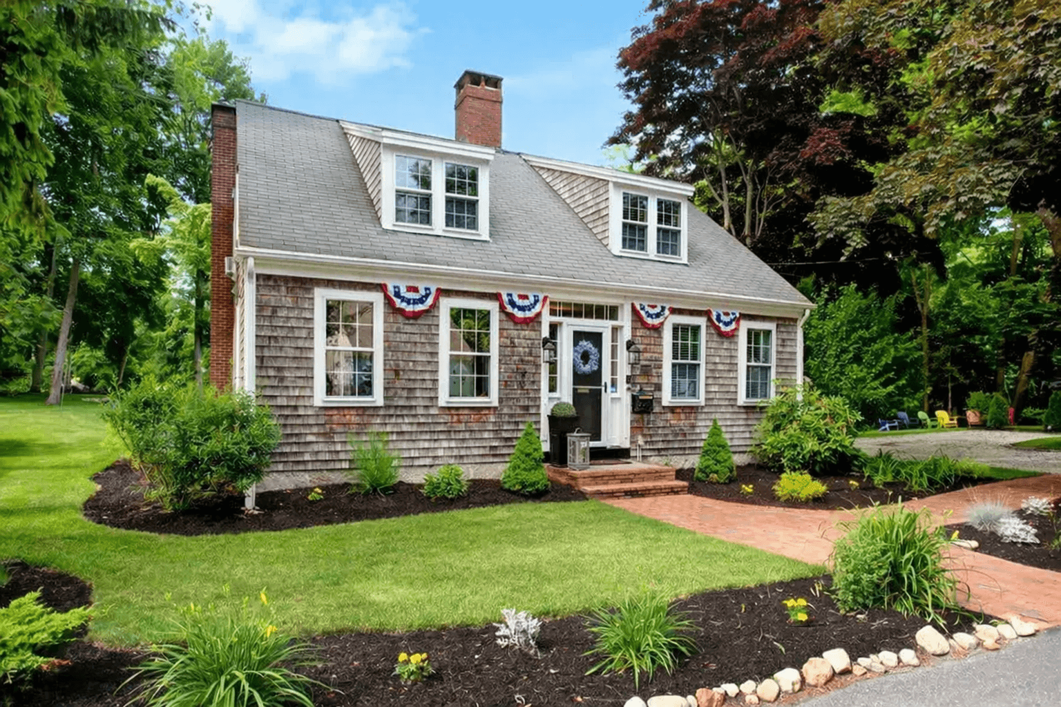 A charming two-story house with a shingle exterior, decorated with patriotic bunting, surrounded by a lush garden.
