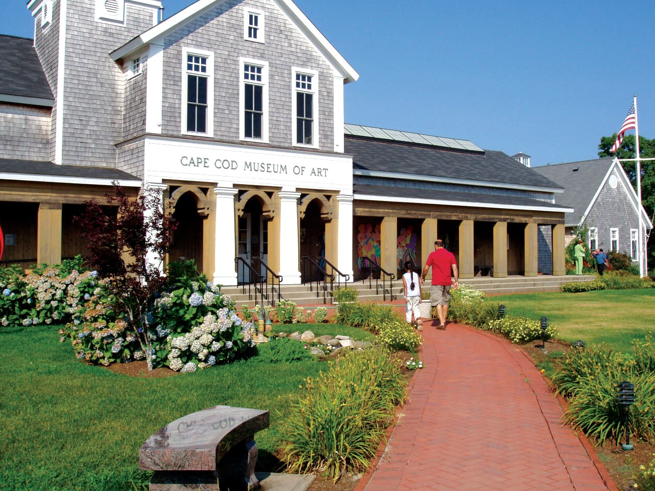 A couple walks along a path towards the Cape Cod Museum of Art, surrounded by greenery and flowers.