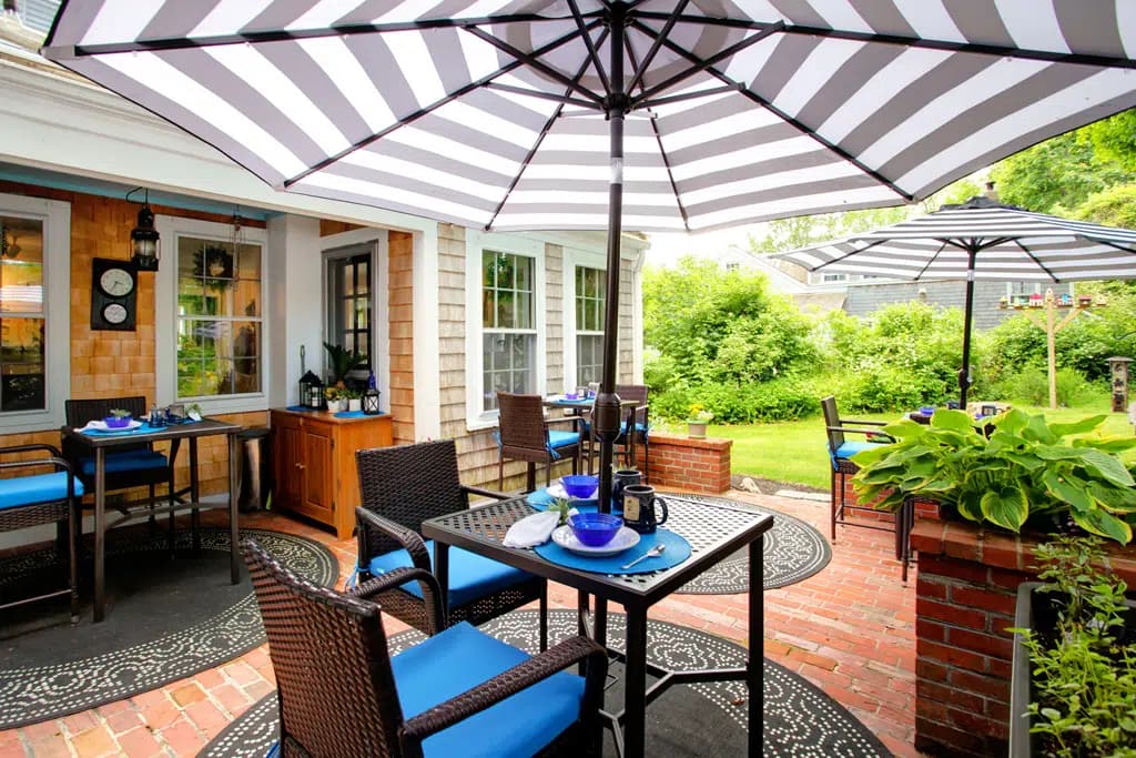 Brick patio, square black tables, round black and white rugs with patio chairs and lime green cushions with a backdrop of the cedar shake building.
