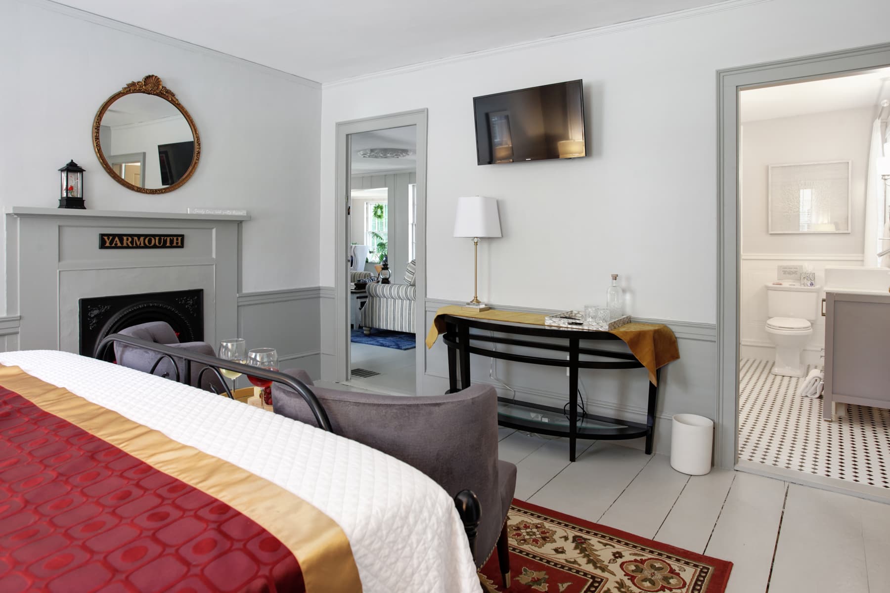 View from bed showing back of two gray chairs, decorative fireplace with mirror, side table against far wall, and doorway leading into bathroom