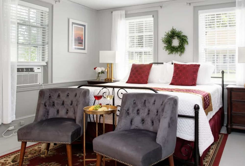 Dark iron bed, white linens, burgundy pillows, nightstand and lamp and two grey padded chairs are foot of bed with table.