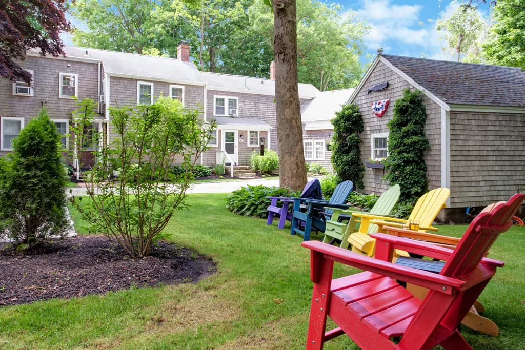 View of side of grey clapboard 2 story inn with multi color adirondack chairs