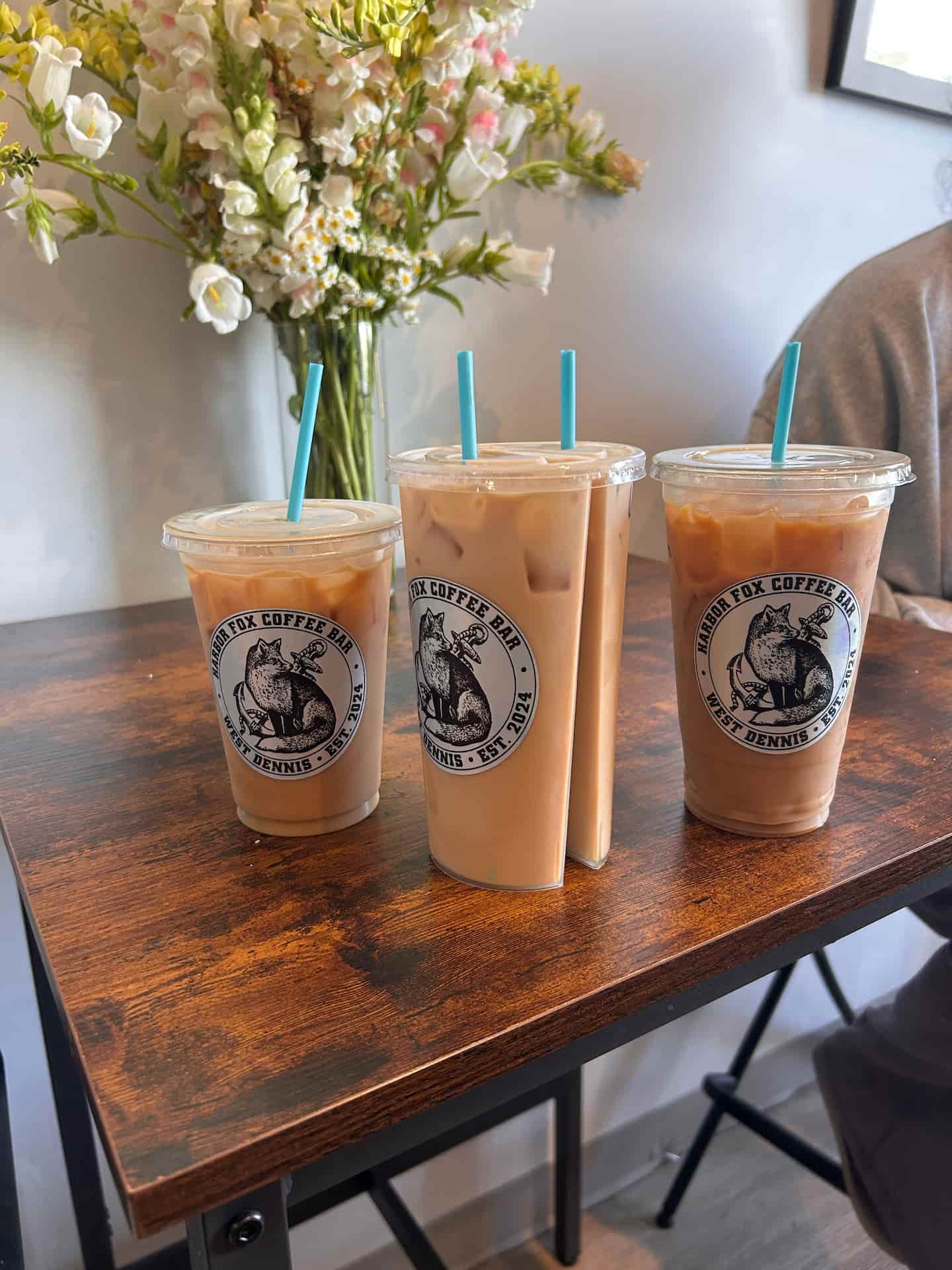 Three iced coffee drinks with teal straws sit on a wooden table, accompanied by a vase of flowers in the background.