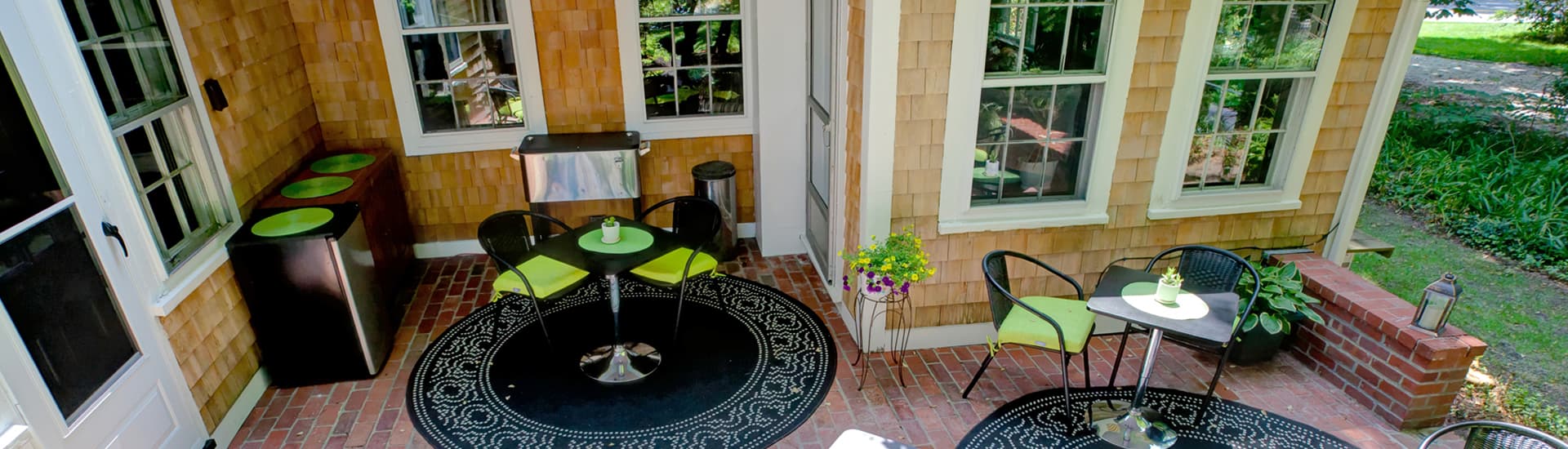 Brick patio, square black tables, round black and white rugs with patio chairs and lime green cushions with a backdrop of the cedar shake building.