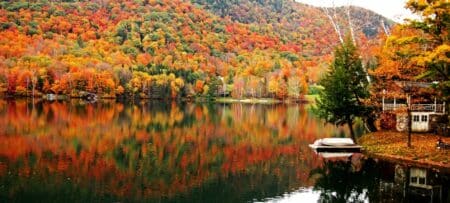 A hill covered with trees with colorful leaves of orange, red, and yellow. Foliage reflects on the water of a lake in the foreground.